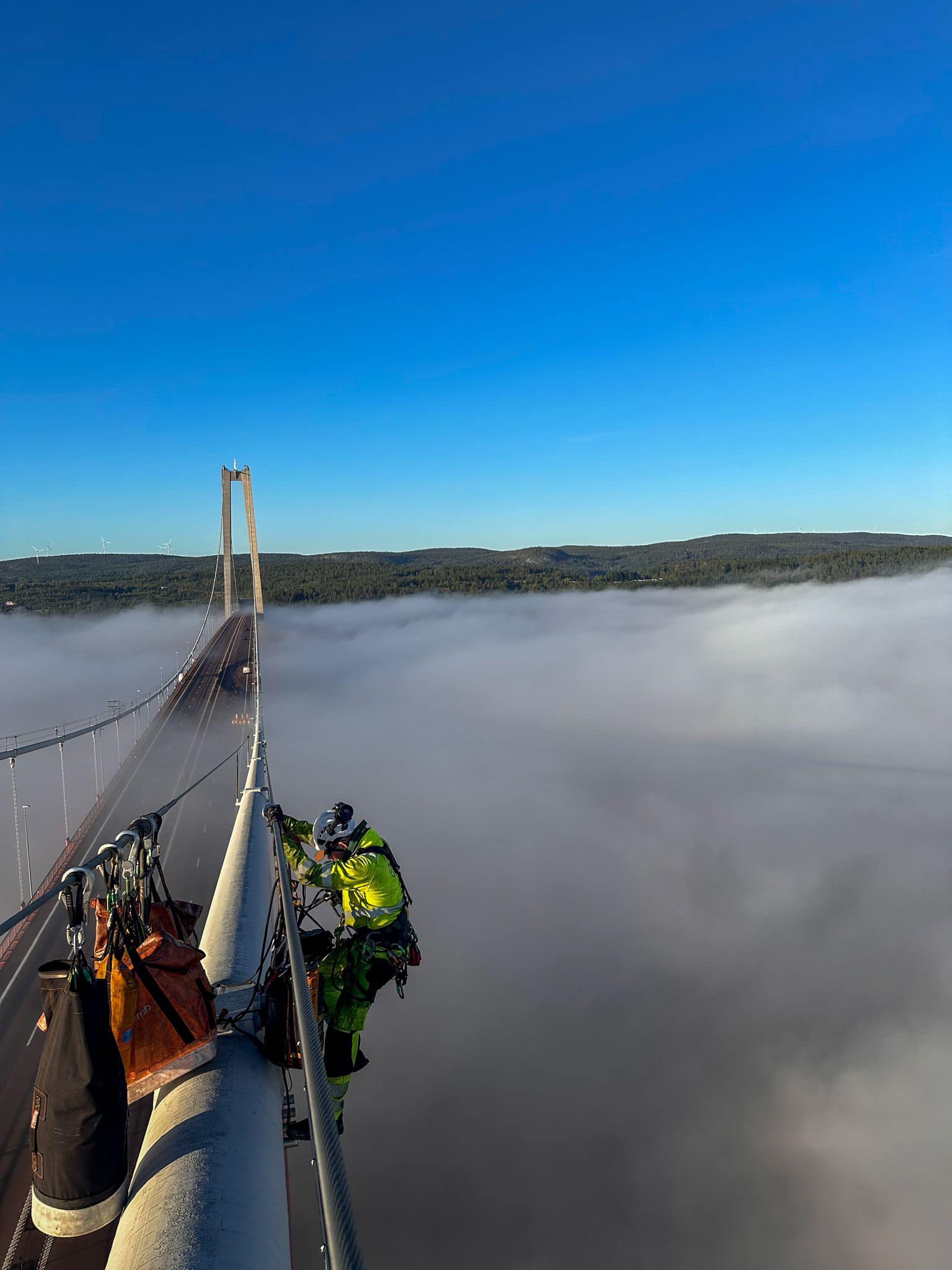 Rope access technician working on a suspension bridge cable above the fog
